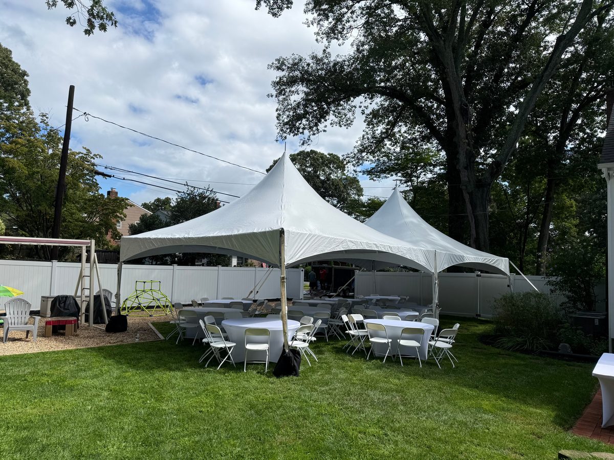 Two white high peak tents with covered round tables and white chairs under a big shade tree